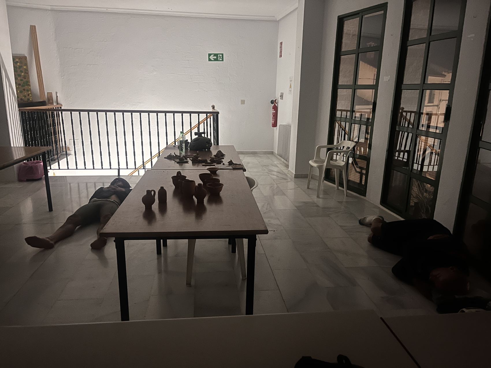 Riders asleep on the tiled floor of a bar at night, around a table of pottery, under a lit emergency exit sign.