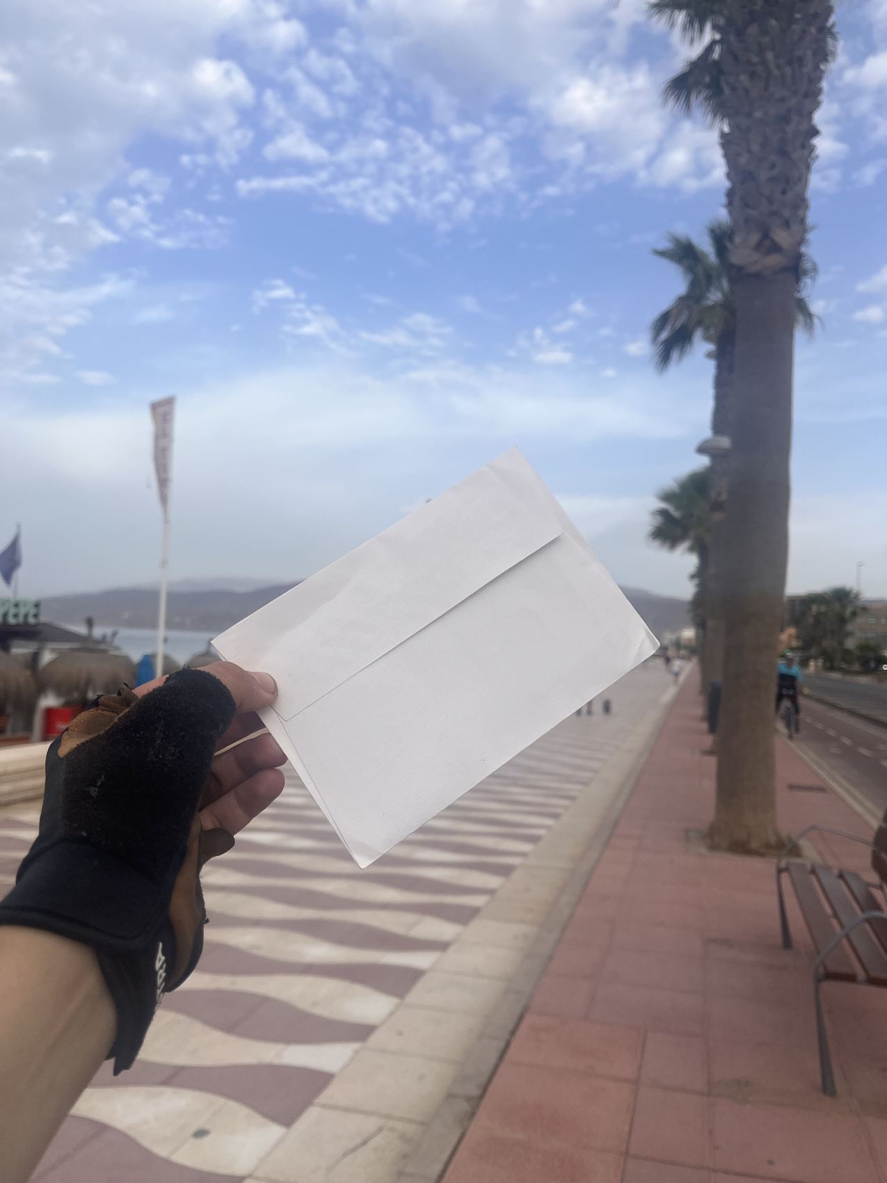 A gloved hand holding the last white envelope up on the Almería seafront, palm trees and the wave-patterned promenade behind.